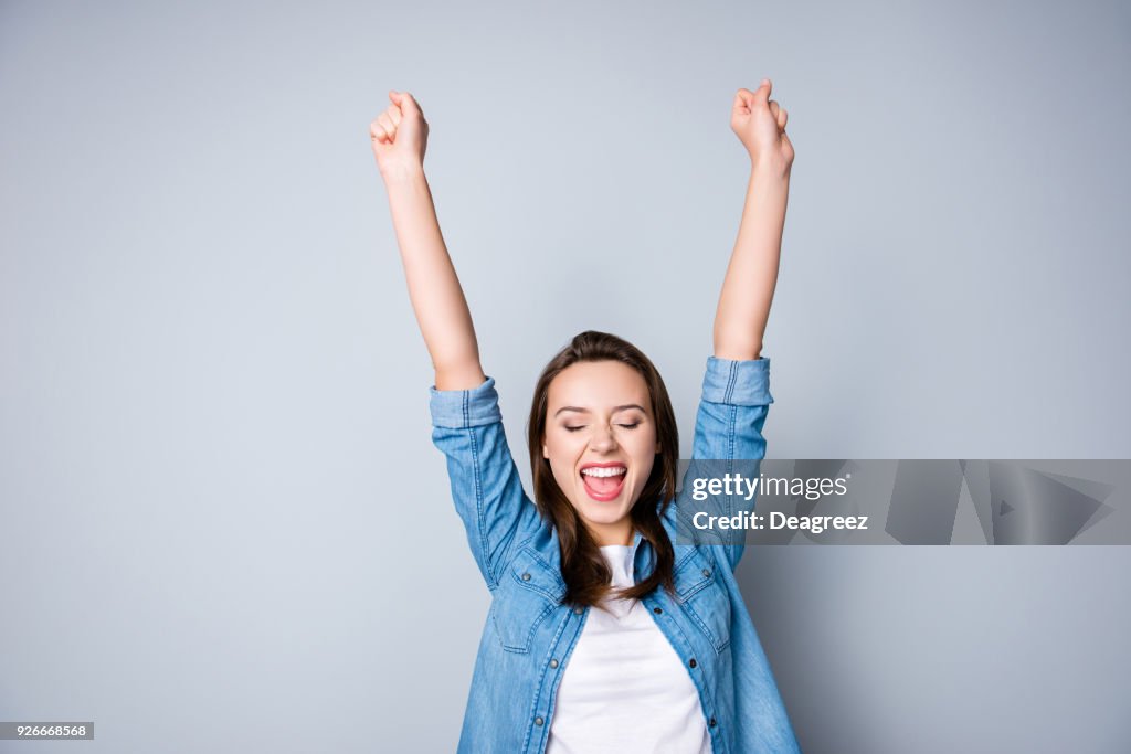 Sorprendida morena joven empresaria en camisa casual es victoria gesticular con las manos levantadas, es sorprendido, muy feliz, con los ojos cerrados, sonrisa, boca abierta en el fondo gris de barrado