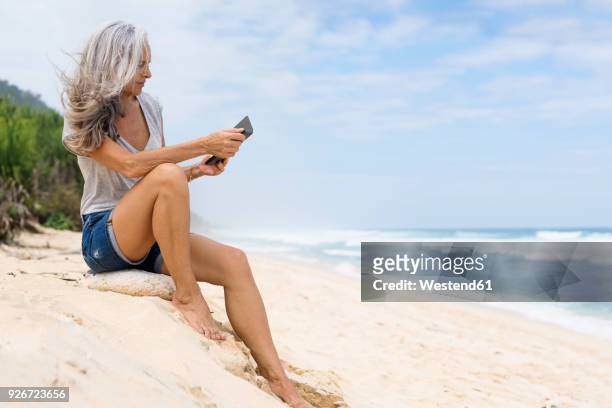 beautiful smiling senior woman sitting at the beach, holding e-book - e reader stock-fotos und bilder