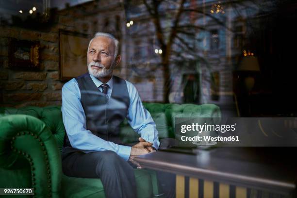 elegant senior man sitting on couch in a cafe looking out of window - goed gekleed stockfoto's en -beelden