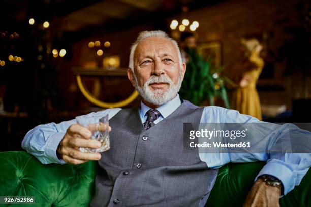 portrait of elegant senior man sitting on couch in a bar holding tumbler - casual businessman glasses white shirt stock pictures, royalty-free photos & images