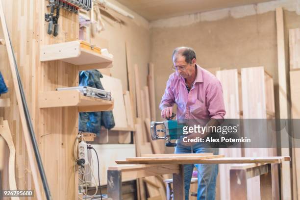 o homem de 65 anos de idade sênior trabalhando com a máquina de trituração de mão elétrico. tratamento de madeira na fábrica de móveis pequenos. - 65 69 years - fotografias e filmes do acervo