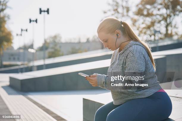 curvaceous young woman training, sitting on wall looking at smartphone - buxom blonde fotografías e imágenes de stock