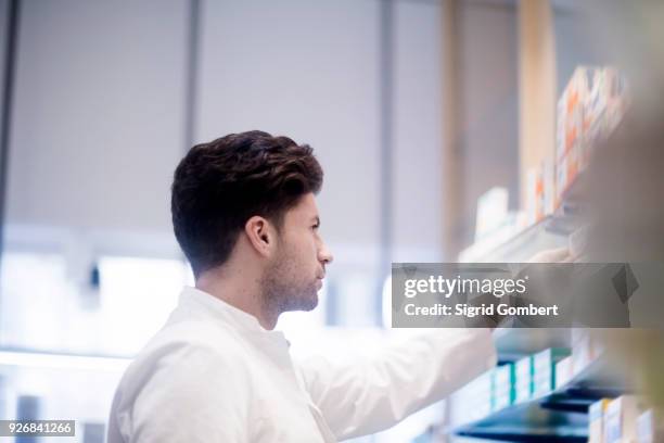 young male pharmacist removing medical drug from pharmacy shelves - sigrid gombert - fotografias e filmes do acervo