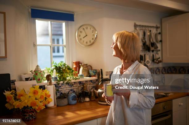 senior woman with hot drink looking out of kitchen window. - old clock stock-fotos und bilder