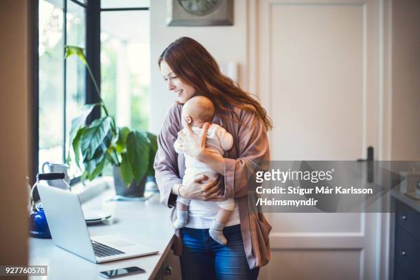 mother using laptop while carrying baby in kitchen - mother and baby and laptop stock pictures, royalty-free photos & images