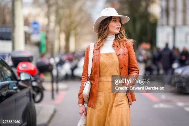 Angelica Ardasheva wears a white hat, a brown leather jacket, orange dress, brown leather boots, outside Giambattista Valli, during Paris Fashion...