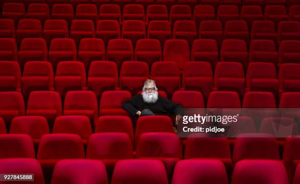 een man met witte baard zit in een lege bioscoop of theater met comfortabele rode stoelen - cinema seats stockfoto's en -beelden