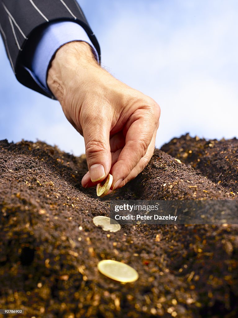 Businessman's hand planting gold coins in soil.