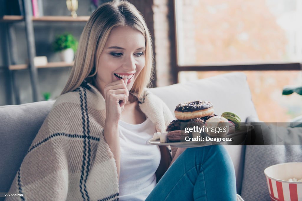 Portrait of beautiful emotional charming attractive sweet toothy woman sitting on sofa in living room, holding plate of donuts and macaroons, looking exciting satisfied