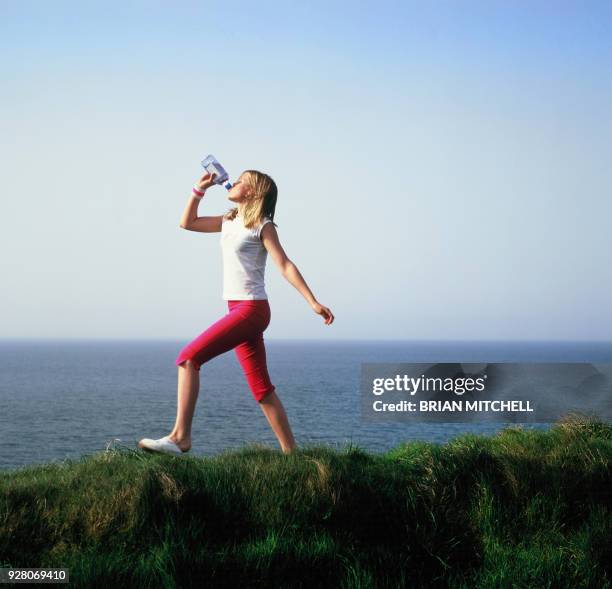 blond teenage girl in summer sports clothing exercising and walking a coastal foot path, drinking bottled water - women in wet tee shirts stock pictures, royalty-free photos & images