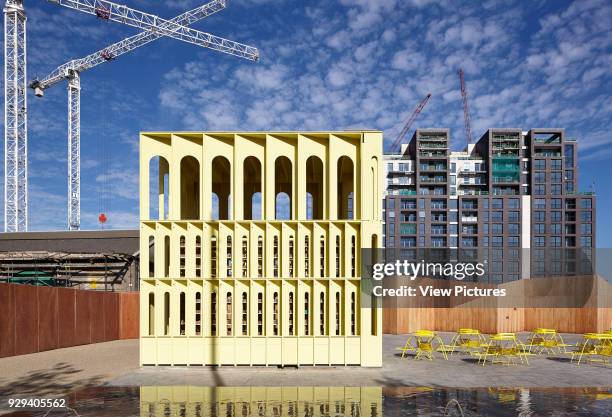 Pavilion with cranes and construction behind, reflections in the water of the fountain below. Yellow Pavilion, London, United Kingdom. Architect:...