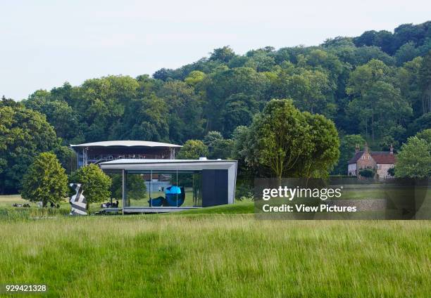 Distant elevation of glass pavilion with Jeff Koons sculpture and Wormsley estate. Island Pavilion and Footbridge, High Wycombe, United Kingdom....