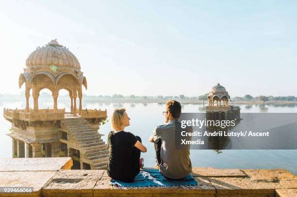 young couple look out at gadi sagar lake in morning light - indian couple foto e immagini stock