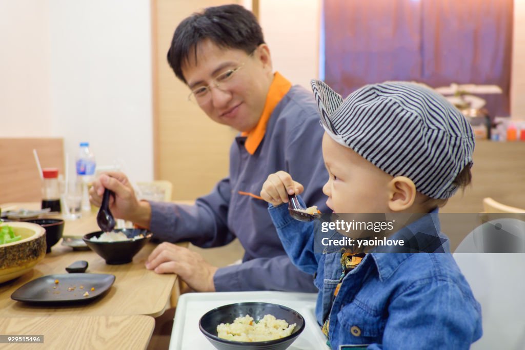 Cute Asian 18 months toddler baby boy child eating food with fork & spoon by himself at Japanese restaurant, Dad proud of hime