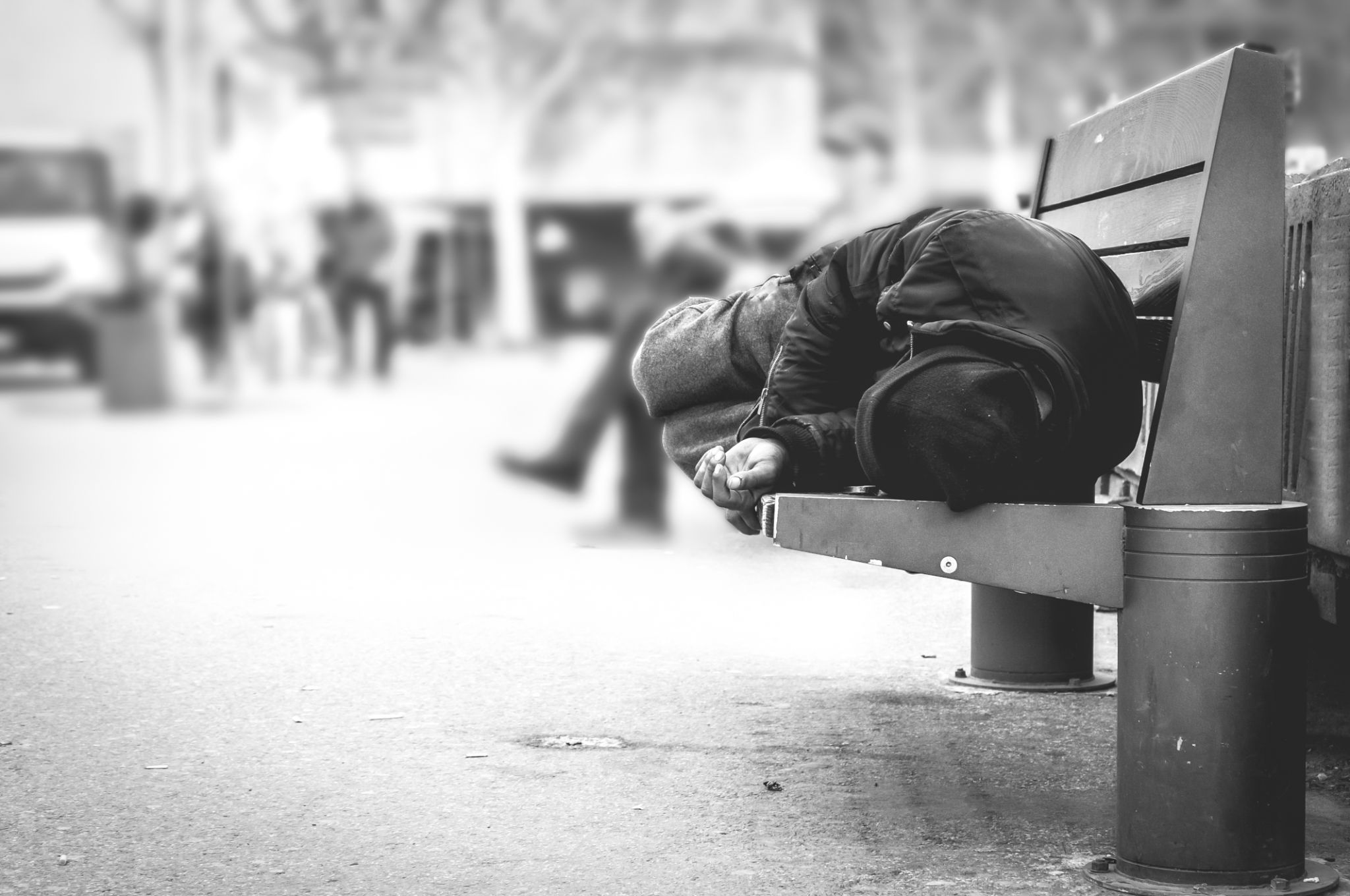 Poor homeless man or refugee sleeping on the wooden bench on the urban street in the city, social documentary concept, selective focus, black and white Poor homeless man or refugee sleeping on the wooden bench on the urban street in the city, social documentary concept, selective focus, black and white