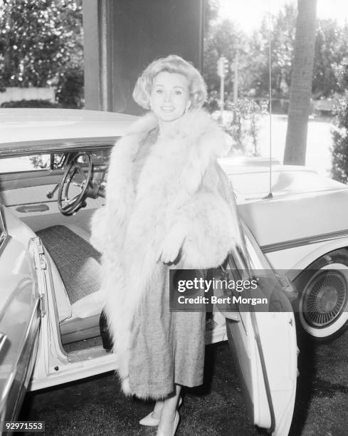 Zsa Zsa Gabor standing beside an open car door wearing a fur trimmed coat, Palm Beach, Florida, 1957