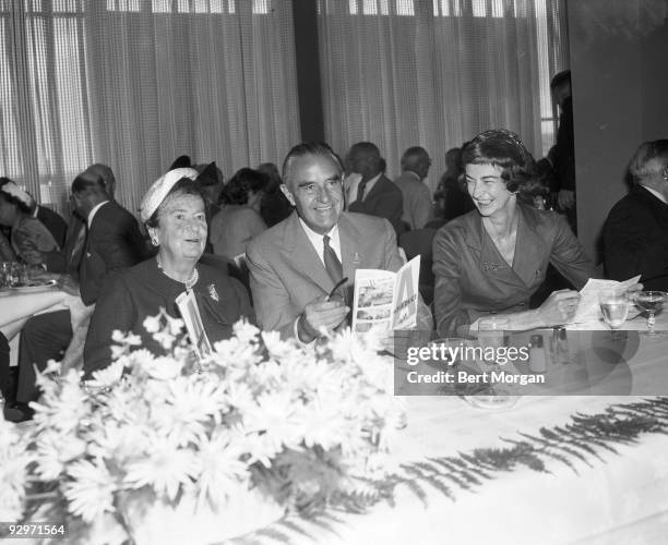 Mrs George D Widener, Hon William Averill Harriman, and Mrs Edward T Dickinson laughing while they sit at a banquet table holding racing forms on...