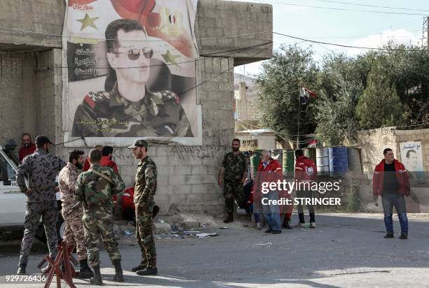 Picture taken on March 9, 2018 shows members of the Syrian government forces and the Syrian Red Crescent waiting by a poster depicting President...