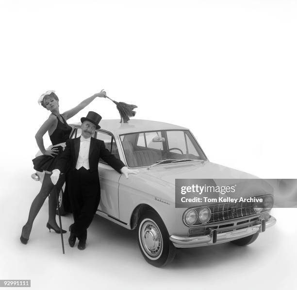 Man dressed in a formal tuxedo, top hat, and holding a walking stick, stands next to his Datsun automobile while a taller woman dressed as a French...