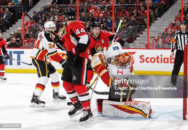 David Rittich of the Calgary Flames has the puck go off his face-mask as Jean-Gabriel Pageau and Mark Stone of the Ottawa Senators look for the...