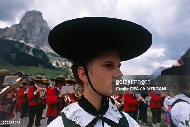 Woman wearing a hat and traditional clothes, with a marching band in the background, Ladin country festival, Corvara, Val Badia, Trentino-Alto Adige,...