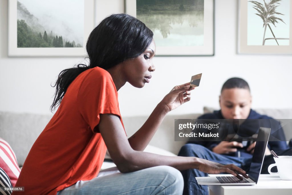 Mother holding credit card while using laptop with son in background at home