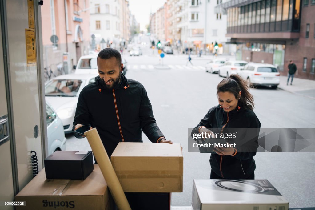 Smiling coworkers using technologies while examining packages in delivery van