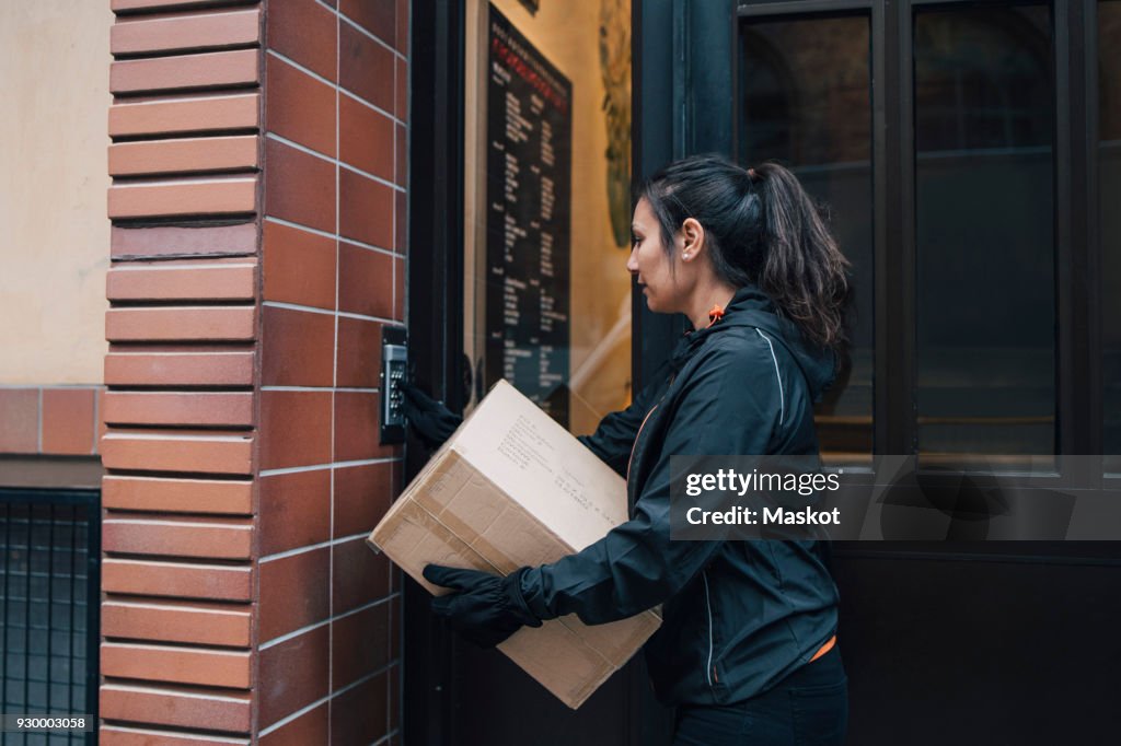 Female messenger ringing intercom while carrying box by closed door