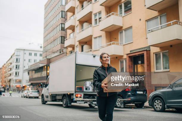 smiling female messenger carrying box while walking in city - pakket stockfoto's en -beelden