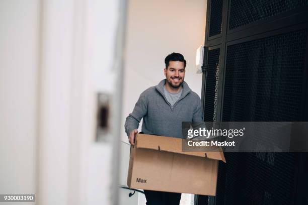 smiling man carrying cardboard box while walking by metal grate in house - box white flat imagens e fotografias de stock