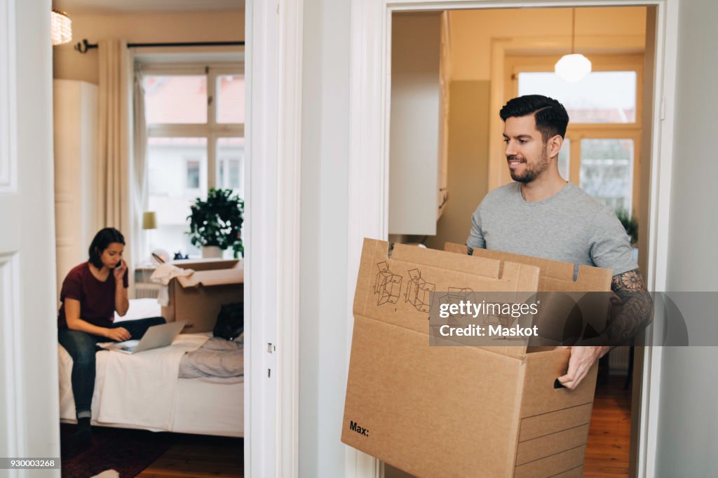 Smiling man carrying box from doorway towards woman using laptop in bedroom