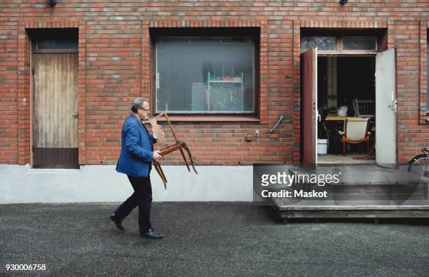 full length of senior man carrying wooden chair while walking by workshop - side view carrying stock pictures, royalty-free photos & images