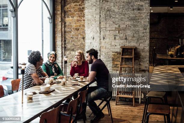 cuatro amigos sentado en una mesa larga de café ventana hablando - café bar fotografías e imágenes de stock