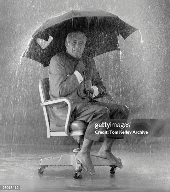 Studio portrait of a shoeless businessman sits on an office chair, while holding a broken umbrella above him as rain crashes all around, 1998.