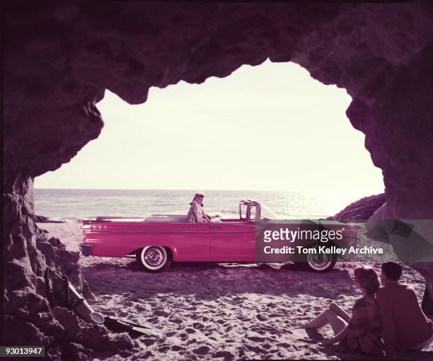 View of parents sitting inside a beach cave while watching their daughter who sits on their red Mercury Monterey convertible, 1959.