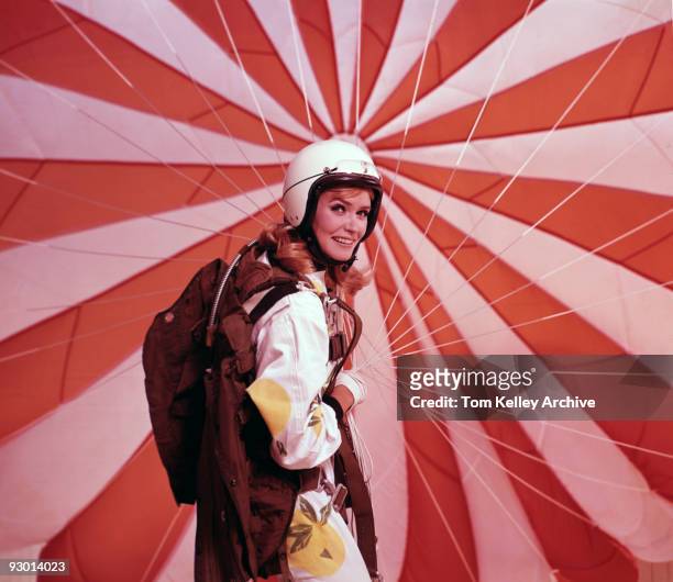 Studio depiction of a woman looking back while in the process of folding up her red and white striped parachute after landing from a jump, 1966.