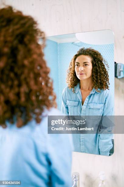 curly haired woman stands in the bathroom looking at her reflection in the mirror - woman in mirror stock pictures, royalty-free photos & images