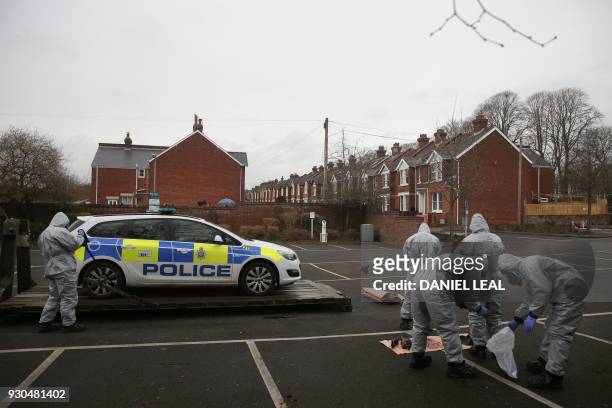 Military personnel wearing protective coveralls load a police car onto a military vehicle for it to be taken away from a cordoned off area behind a...