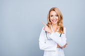 Look there! Portrait of confident experienced qualified cheerful smiling with blonde hair mature female doc, she is pointing on blank empty space over her shoulder, isolated on grey background