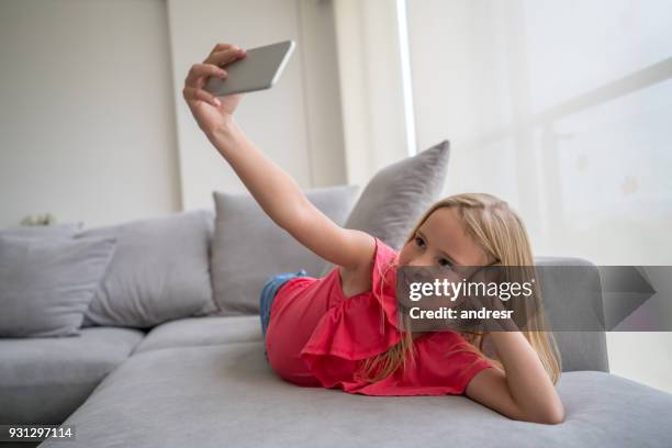 beautiful blond little girl at home taking a selfie with a smartphone while lying down on the couch looking happy - girl lying down stock pictures, royalty-free photos & images