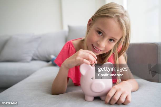 süßes kleines mädchen münzeinwurf in ihr sparschwein, blick auf die kamera zu lächeln - blonde girl piggy bank stock-fotos und bilder