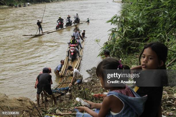 Two girls sit on the river bank as several people assist motorcyclists to pull up their vehicles onto wooden raft to cross Cisadane River, in Rumpin,...