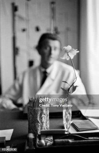 View of a cup of pencils and a single rose in a vase on a desk as French fashion designer Hubert de Givenchy sits in the background, Paris, France,...