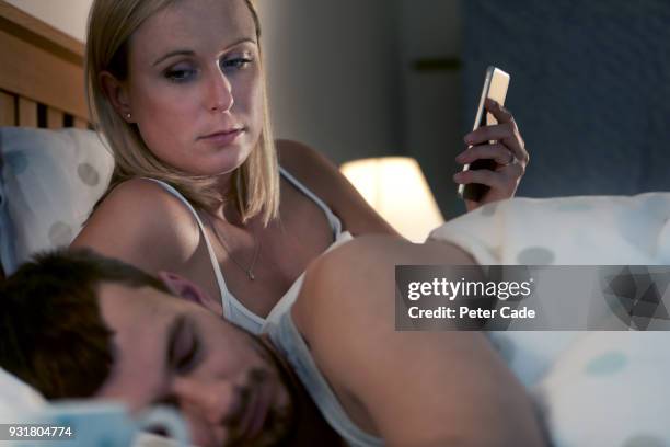 woman on phone in bed next to sleeping partner - verraad stockfoto's en -beelden