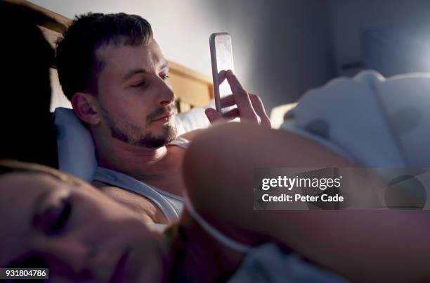 man on his phone in bed next to sleeping partner - mente foto e immagini stock