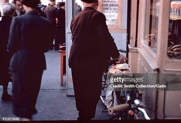 Close-up of a French man leaving a boulangerie in Paris, France, with three loaves of baguette bread strapped to the back of his bicycle, 1955.