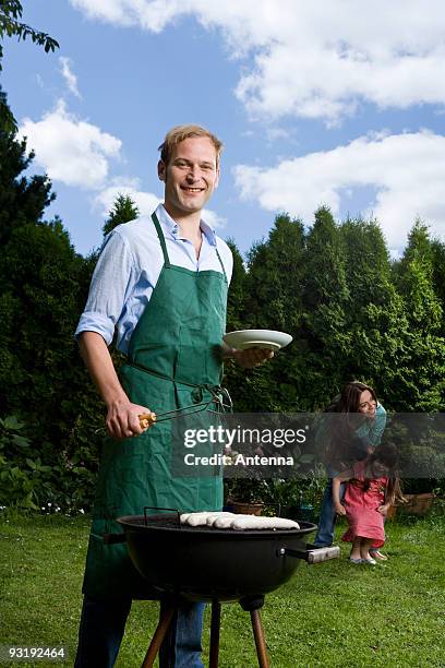 a man barbecuing bratwurst in a back yard, wife and child in the background - apron man stock pictures, royalty-free photos & images