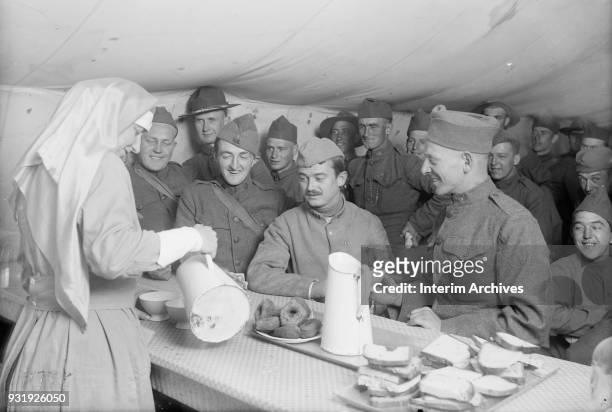 Surrounded by smiling American soldiers, a French soldier watches as an American Red Cross canteen worker pours coffee, Toul, France, June 1918. An...
