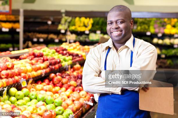 portrait of cheerful male grocer standing in produce section - fruit stand stock pictures, royalty-free photos & images