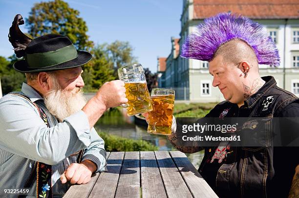 germany, bavaria, upper bavaria, man with mohawk hairstyle and bavarian man holding beer stein glasses - duality stock-fotos und bilder